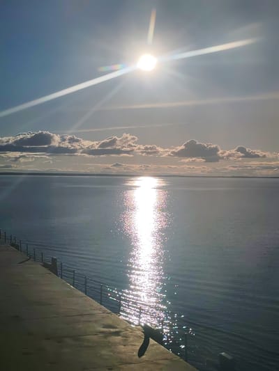 The image depicts a bright, sunny day over a body of water. The sun is high in the sky, producing significant glare and reflection on the water surface. Some clouds are visible on the horizon. In the foreground, there is a concrete walkway or pier with a metal railing. The location is possibly in Kostyaevka, Russia. No people are present.