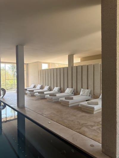 The image depicts a serene indoor spa area with a swimming pool. Several white lounge chairs, each with a white pillow and a rolled towel, are neatly arranged alongside the pool. The walls are textured in a light beige, and a textured partition with vertical grooves divides the lounging area. Large windows offer a view of lush greenery and trees outside. The swimming pool itself is dark and appears to be illuminated by underwater lights. The overall ambiance is one of tranquility and modern, minimalist design. This scene is located in the city of Artà, Spain.