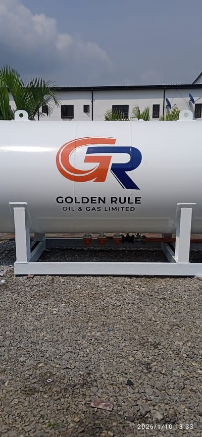 The image displays a large, white, horizontal cylindrical storage tank positioned outdoors in Isheri-North, Nigeria. The tank features a prominent logo with an intertwined orange 'G' and blue 'R', beneath which "GOLDEN RULE OIL & GAS LIMITED" is printed in black uppercase text. The tank is supported by two white, rectangular-frame structures. Below the tank, several orange-capped components are connected to a reddish-brown pipe. The ground consists of grey gravel, with scattered small debris including a red rectangular fragment and a white ring. In the background, a light-colored building with multiple dark rectangular windows and a dark roofline is partially visible, partially obscured by green palm fronds. Two blue satellite dishes are mounted on the right side of the building's roof area. The sky above is blue with scattered white clouds. No individuals are present in the scene. A digital timestamp "2026/1/10 13:33" is visible in the lower right corner.