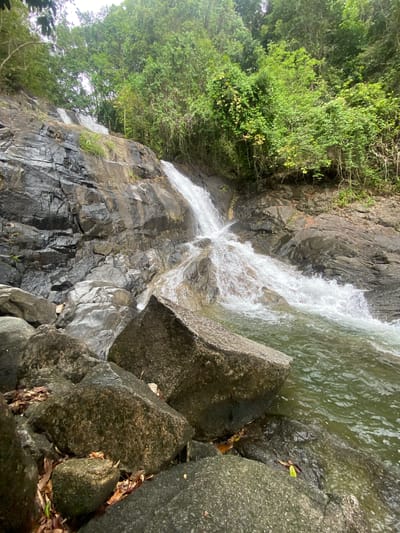 The image shows a waterfall flowing over rocks in Thai Mueang, Thailand. The water is clear and cascades down a dark, rocky cliff face. Lush green vegetation covers the top left of the frame. Large, moss-covered boulders are situated at the base of the waterfall, to the right of the frame, interspersed with some leaf litter.