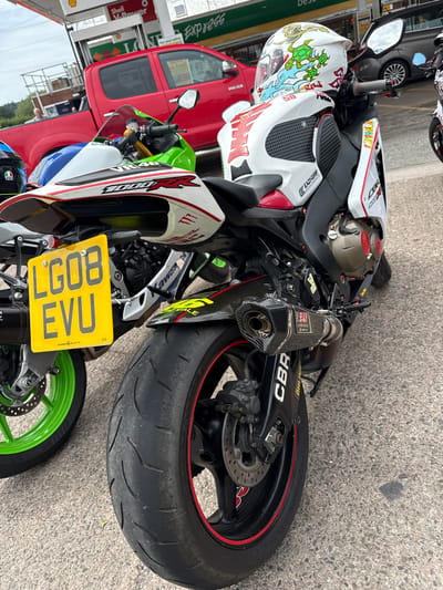 The image shows a close-up view of two motorcycles parked on a paved surface, likely at a gas station, in Much Dewchurch, United Kingdom. The motorcycle in the foreground is a white and red sports bike, identified by the "CBR" branding and "1000RR" on its fairing. It features a yellow license plate with the registration "LG08 EVU". A white helmet with colorful graphics, including turtles and sharks, is placed on the seat. The motorcycle has a carbon fiber exhaust system. Behind it, a portion of a lime green motorcycle with a blue and white fairing is visible, as is a red pickup truck. The background includes a Shell V-Power gas pump.