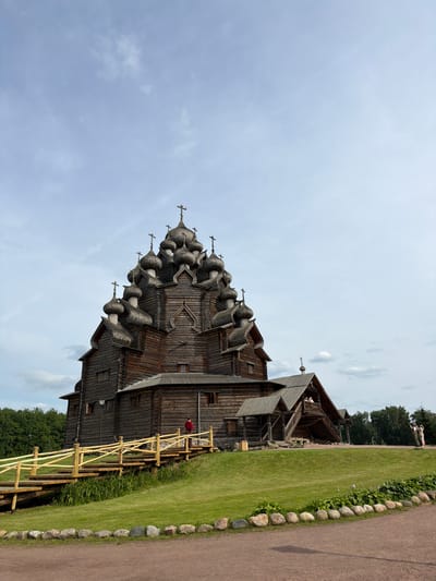 This is a wide-angle, low-angle outdoor photograph of the Church of the Transfiguration in Severnaya Samarka, Russia. The large, wooden church is the dominant subject, characterized by its dark timber construction and numerous onion domes topped with crosses. The church is situated on a grassy hill, with a wooden walkway leading up to its entrance. A single person in a red jacket stands on the walkway. To the right of the church, two people are visible on an outdoor staircase, and another person is taking a photo with a lamp. The sky is a pale blue with scattered white clouds. The foreground consists of a dirt path bordered by rocks and a strip of grass with some plants. Trees line the background.