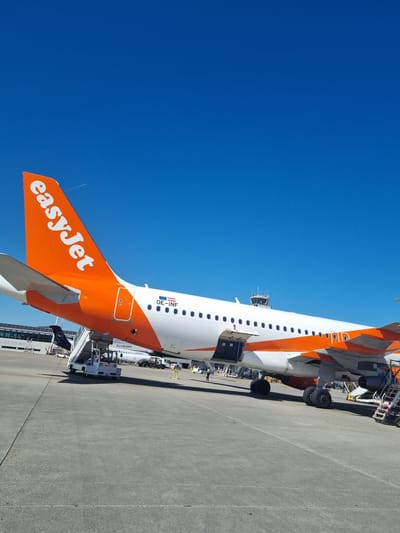 The image shows an EasyJet Airbus A320 aircraft parked at EuroAirport Basel-Mulhouse-Freiburg, located in Saint-Louis, France. The plane's tail, painted in EasyJet's signature orange, with the company logo, is prominently displayed against a clear blue sky. The aircraft is on the tarmac and ground support equipment is positioned adjacent to the plane, including a baggage loader at the cargo hold and passenger stairs at the front. The plane is mostly white with orange accents around the front and rear of the fuselage. The aircraft's registration number, OE-INF, is visible near the tail. An airport terminal building is visible in the background.