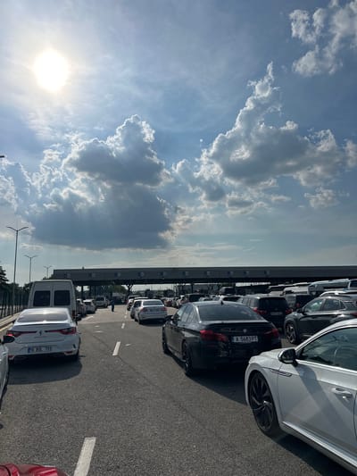 The image displays a bustling daytime scene at the Kapitan Andreevo border crossing into Bulgaria, characterized by a long queue of vehicles under a partly cloudy sky. The perspective is from inside a red vehicle situated in one of the multi-lane approaches to the border checkpoint.
The upper portion of the image shows a bright blue sky dominated by scattered cumulus clouds, some casting visible crepuscular rays. The sun is prominent in the upper left, emitting a strong glare and lens flare, indicative of a high sun angle.
The lower half captures the asphalt road, segmenting into multiple lanes packed with queued cars. A large, grey overhead structure spans across these lanes, featuring "РЕПУБЛИКА БЪЛГАРИЯ" and "REPUBLIC OF BULGARIA" in white lettering, confirming the border entry point. Below this main sign, several smaller panels or electronic displays are visible. On the left side of the road, two streetlights on poles and a metal fence run parallel to the traffic.
Vehicles of various makes, models, and colors are densely packed in the queues. In the leftmost visible lane, a white Renault Megane sedan (Turkish plate: 16 BUL 701) is followed by a white commercial van. In the central lane, a black BMW 5-series sedan (Bulgarian plate: A 5683 PT) is in the immediate foreground, followed by a dark SUV (German plate: M:VV 4841). Further down this lane, a single individual stands beside a vehicle. On the right, partially visible, is a white Volkswagen Arteon, followed by a dark SUV and a white sedan (Turkish plate: 32 EJ 800). Numerous other cars and vans extend into the background across all lanes, suggesting significant traffic congestion.
The overall environment is an open-air border facility, with minimal surrounding foliage immediately adjacent to the road, beyond the fence. The primary activity depicted is the slow, stationary queueing of vehicles awaiting processing at the border.
