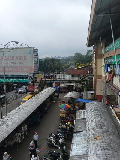 An elevated shot captures a bustling outdoor market adjacent to a building with green signage. Rows of stalls with metal roofing line the walkway, hosting vendors and shoppers. Parked motorcycles fill the space. Above the market, a pedestrian bridge crosses the street lined with traffic. A large billboard advertises available space with a phone number. The sky is overcast.