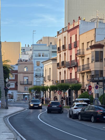The image shows a street scene in Vinaròs, Spain. Buildings of varying heights and colors line the left side of the street. Cars are parked along the right side of the street, which has a winding road marked by white lines. A black car is approaching the camera, while another is parked ahead of it. Pedestrians are visible near the parked cars. Street signs and trees are positioned along the road. The sky is blue with few clouds.