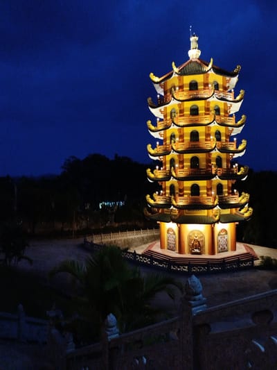 Captured at night, the image features a tall, illuminated pagoda in Phú An, Vietnam. The pagoda, likely of Buddhist or Taoist origin, has multiple tiers with curved roofs and decorative elements. It is brightly lit with warm-toned lights, contrasting against the dark blue night sky. Below the pagoda, a walled walkway and hints of foliage are visible. A stone railing and some greenery are also visible in the foreground, suggesting the vantage point is elevated. The background is a blend of dark trees and vague structures.
