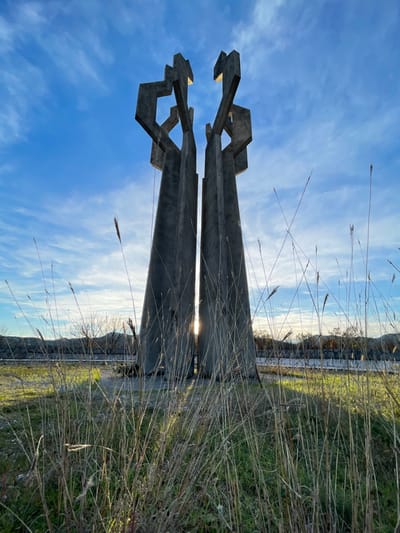 The image displays the brutalist concrete "Memorial to the Fallen Fighters" monument located in Barutana, Montenegro. The monument consists of two tall, tapering concrete structures, each with geometric, angular extensions at its apex, resembling abstract figures or forms. The sun is positioned directly between these two main structures, creating a prominent lens flare and backlighting the monument, with long shadows cast towards the viewer, indicative of either sunrise or sunset. The immediate foreground features a dense field of tall, dry, straw-colored grasses interspersed with patches of green foliage. In the midground, the base of the monument sits on a gravelly or dirt patch. The background reveals a clear blue sky with scattered thin, white cirrus clouds, a visible horizon line, bare trees and shrubs on the left, and distant hills or mountains silhouetted against the bright sky. No individuals, vehicles, or active human interactions are present within the scene.