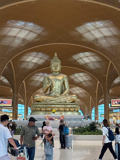 In the city of Kandal Stung, Cambodia, a large golden Buddha statue sits centrally within a vast, open-air building, likely an airport terminal, characterized by its high, arched ceiling with a latticed wooden design and geometric light fixtures. The statue, seated on a tiered platform adorned with decorative motifs, is bathed in light, highlighting its intricate details and serene expression. Surrounding the base of the statue are planters filled with lush green foliage and scattered floral arrangements.
In the foreground, a group of people navigate the spacious, polished floor. To the left, a person in a white shirt pushes a luggage cart. Near the center, a man wearing a green short-sleeved shirt and a dark baseball cap holds a mobile phone in his hand, facing away from the camera. Beside him, a young child in a denim outfit with arms raised walks towards the right. To the right of the child, a woman with blonde hair, wearing a pink top, looks at her phone. Further to the right, a woman with dark hair and a backpack walks away from the camera. The background features large windows revealing a blue sky and architectural elements of the building, including digital display screens and other people moving through the space.
