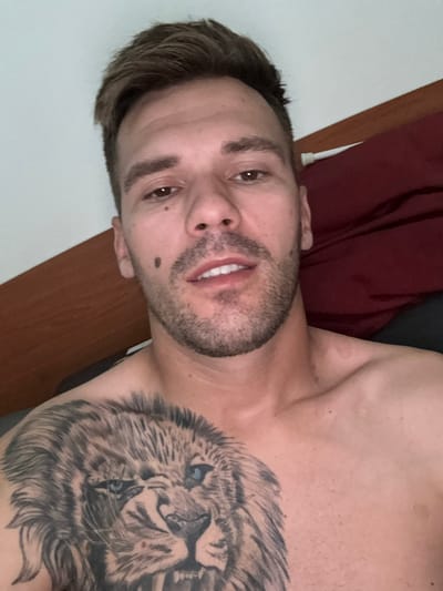 The image shows a close-up, rotated view of a man with a tattoo of a lion on his chest. He has short, dark hair, a beard, and a mole on his left cheek. His eyes are open and he appears to be smiling slightly, with his teeth visible. The man is shirtless, and part of his shoulder and chest are visible. In the background, there is a dark red fabric, possibly clothing or bedding, and a wooden surface that could be furniture. The lighting suggests it is daytime. The man is likely indoors, and the overall impression is casual and relaxed. The location context is Sesto Calende, Italy.
