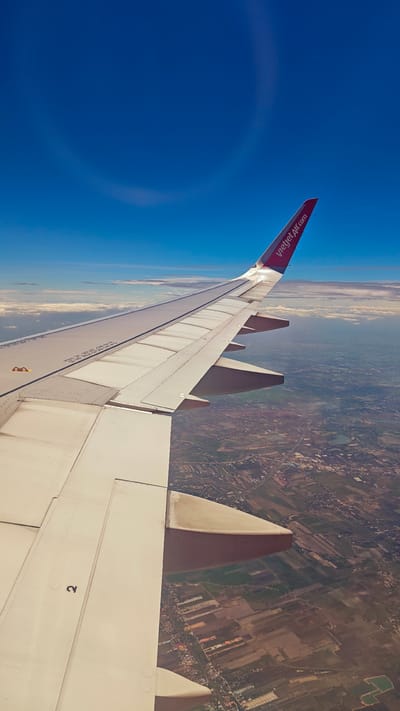 An aerial view from an airplane window shows a landscape below with cultivated fields and scattered buildings, characteristic of the Ban Chang Subdistrict in Thailand. The airplane's wing, painted white with a red tip bearing the "vietjetair.com" logo, dominates the foreground, extending diagonally across the frame. The sky above is a vibrant, clear blue, with a faint circular atmospheric optical phenomenon visible in the upper left quadrant. The ground below is a patchwork of green and brown fields, interspersed with roads and small clusters of structures, indicating a rural or semi-rural setting within the subdistrict.