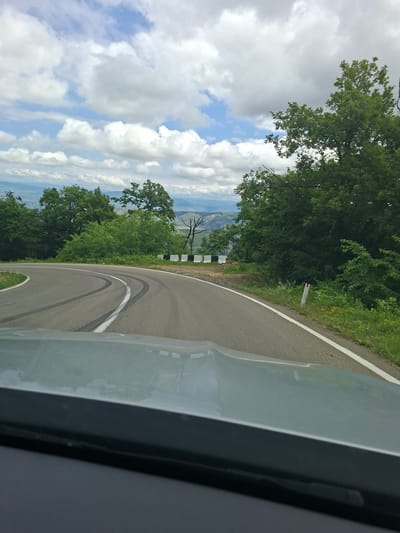 The image shows a view from inside a vehicle looking out at a winding road in a mountainous, forested area. The road has tire marks indicating recent sharp turns. A concrete barrier with black and white markings lines the side of the road. Lush green trees and vegetation are abundant on either side of the road. In the distance, a valley with mountains and a hazy sky can be seen. The sky is partly cloudy with white cumulus clouds against a blue background. A single red and white road marker is visible on the right side of the road. The foreground of the image is dominated by the hood of the vehicle, which is a light gray color and reflects the sky and surroundings. The scene appears to be in the vicinity of Kvemo Nichbisi, Georgia, given the natural landscape and road infrastructure typically found in such regions. There are no people visible in the image.