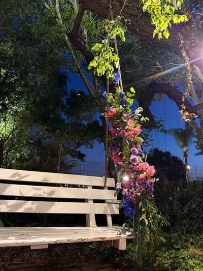 The image captures an outdoor scene at night or late dusk, illuminated by artificial lights. The central focus is an elaborate floral arrangement draped across a rope or twine structure, which appears to be part of an entrance or archway, leading to or from a white wooden gate on the right.
The floral arrangement is rich with vibrant colors, featuring clusters of pink, fuchsia, purple, and blue flowers, interspersed with various green foliage. Some prominent flowers include what look like pink lilies or alstroemeria, purple stock or delphinium, and blue delphinium. The flowers are lush and artfully arranged, suggesting a festive or ceremonial occasion. Small lights are integrated into or near the floral display, casting bright glows and lens flares in the upper left portion of the image.
The background reveals a deep blue night sky visible through the silhouettes of several large trees with substantial trunks and branches. Green leaves, some brightly illuminated by the lights, frame the upper and lower sections of the image, contributing to an organic, garden-like setting. The white wooden gate on the right side is partially visible, showing vertical slats and hinges, with shadows of leaves cast upon it. No people or visible text are present in the image. The overall atmosphere is serene and celebratory, indicative of an outdoor event or decorated space in Nir Eliyahu, Israel.