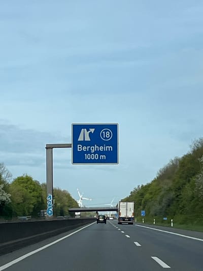 The image depicts a highway scene with a blue road sign indicating the direction to Bergheim, Germany. The sign reads "Bergheim" and "1000 m" and includes the number "18" inside a circle. The sign is mounted on a gray metal pole. The highway has multiple lanes, marked with white lines. A white truck is visible further down the road, along with some other vehicles. Wind turbines are visible in the background. The sky is overcast, and trees line both sides of the highway. Graffiti is visible at the base of the signpost. The location is likely on a highway leading to Bergheim, Germany, based on the sign's information.