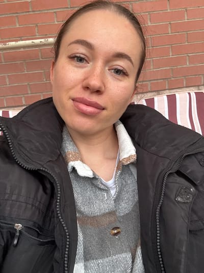 A woman is shown in a close-up shot, wearing a black padded jacket over a grey and white checked shirt, and a white top. The background includes a brick wall and a striped surface. The location is not specified, so it can't be determined that the photograph was taken in Ogryzkovo, Russia.