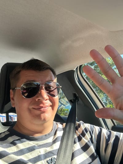 A man with short brown hair and sunglasses is sitting in a car, wearing a blue and white striped t-shirt and a seatbelt. He is smiling and waving at the camera with his right hand. The interior of the car is visible, with the ceiling, seat, and a portion of the window showing trees and the sky outside. The lighting suggests it is daytime. The image does not contain any information that would allow for its location to be identified as Provodina, Montenegro.
