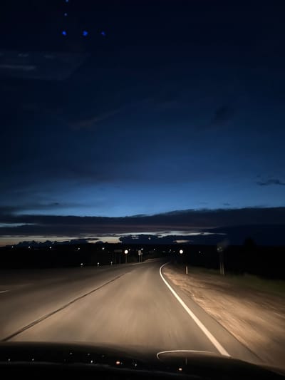 The image captures a view from a vehicle traveling on a paved road at dusk, with headlights illuminating the asphalt. The road curves to the right, bordered by a dirt shoulder. In the distance, a dark silhouette of a treeline is visible, with scattered lights indicating a settlement. The sky is a deep blue, transitioning to lighter shades near the horizon, with streaks of dark clouds. The bottom of the image shows the dashboard and hood of the vehicle. The scene is set in the city of Sars, Russia.