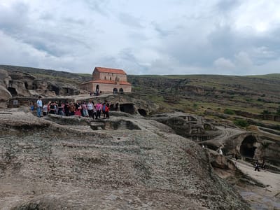 The image depicts the ancient cave city of Uplistsikhe, Georgia, under a cloudy sky. In the mid-ground, a red-tiled roof church with stone walls stands prominently on a rocky outcrop. A group of tourists, dressed in casual clothing, are gathered in front of the church and on the surrounding rocky terrain, observing the site. The landscape consists of rolling hills with sparse vegetation and distinctive rock formations that have been carved into dwellings and structures over centuries. The foreground is dominated by textured, weathered rocks, some appearing to have mossy patches. Pathways and openings carved into the rock are visible, indicating the layout of the ancient city. Several individuals are seen walking along these paths and exploring the carved-out spaces. The overall scene conveys a sense of historical significance and natural beauty.