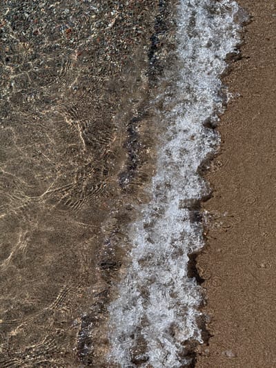 The image is a top-down view of a shallow body of water meeting a sandy and pebbly shore. The water is clear, revealing the textured bottom of small stones and sand. Ripples and reflections create patterns on the water's surface, suggesting movement from a gentle current. A line of foamy white water marks the edge where the water is actively interacting with the shore. The shore itself is a mixture of fine sand and small pebbles, with a slightly darker, wet appearance where the water has receded. The overall impression is a natural, undisturbed scene likely found at the edge of a lake or a slow-moving river. The location is the shore of Lake Issyk-Kul in Koshkol', Kyrgyzstan.