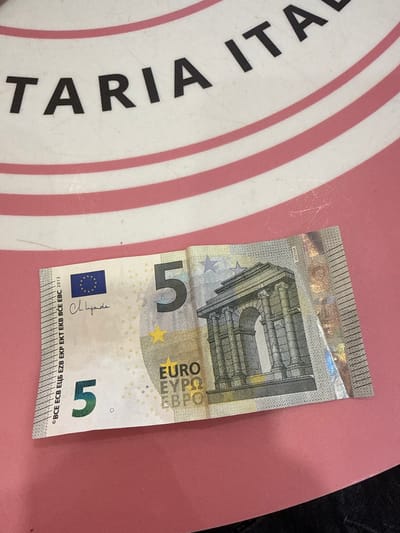 A five euro banknote is shown lying on a pink, circular surface with red stripes. The banknote is oriented diagonally, with the lower left corner closer to the viewer. The front of the banknote is visible, featuring the European Union flag, a signature, and the numeral "5" in large print. The background surface has text that reads "TARIA ITAL". This scene is not specifically identifiable as being in Sesimbra, Portugal.