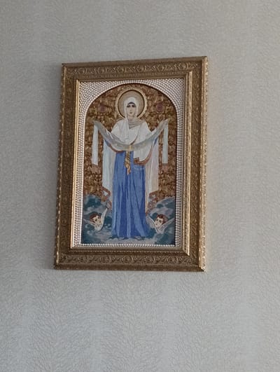 The image shows a framed icon hanging on a textured, light-colored wall. The icon depicts a figure, likely the Virgin Mary, in a blue robe and white headscarf, holding a white veil or cloth. She is surrounded by a golden halo and stands above two small cherub-like figures. The background of the icon features a pattern of flowers and leaves. The frame is ornate and gold. The location of the scene is in the city of Barvinok, Ukraine.