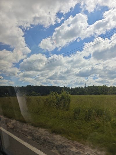 The image depicts a rural landscape, likely taken from a vehicle traveling along a road. In the foreground, a white line marks the edge of the asphalt road, with a grassy shoulder to the right. Beyond the shoulder, a vast, green field stretches towards a dense line of trees in the middle ground. The trees form a dark green, undulating horizon. Above the trees, a bright blue sky is filled with fluffy white cumulus clouds. Several dark, thin wires traverse the sky, suspended between unseen poles. A tall, lattice-style tower, possibly a power pylon, is visible rising above the tree line in the distance. The overall impression is one of a sunny day in the Russian countryside.