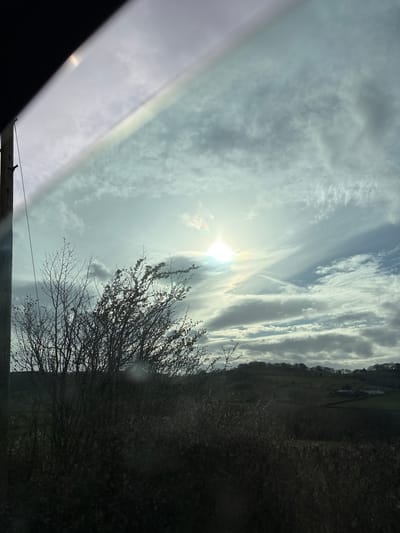The image shows a view of the sky and landscape from a vehicle. The sun is visible through a mix of cirrus and cumuliform clouds. A bare tree is visible in the foreground to the right. Beyond the tree, a dark, vegetated slope rises, potentially part of the Stiperstones area in the United Kingdom. A part of the vehicle's frame and window is visible in the lower left corner. A power line is also visible near the bottom of the frame.
