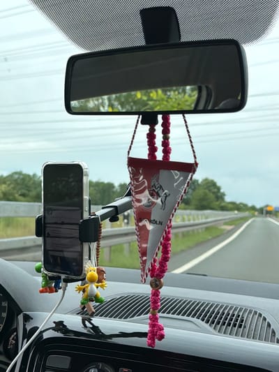 The image is taken from inside a vehicle, likely a car, driving on a road in Langenfeld (Rheinland), Germany. The view is primarily through the windshield, which has a rearview mirror attached. A smartphone is mounted on the dashboard, displaying a dark screen. A small pennant with the emblem of the football club FC Köln hangs from the phone mount. A string of colorful beads and figurines is also attached to the phone. The dashboard and ventilation system of the car are visible. Outside the car, the road stretches ahead with green vegetation along the sides. Overhead power lines are visible above. The image is rotated counter-clockwise.