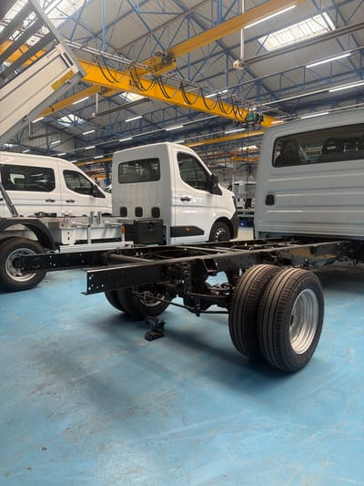 The image shows the interior of a factory in Saint-Berthevin, France. The scene is dominated by the chassis and rear wheels of a light commercial vehicle, likely a truck or van, being assembled. This vehicle is positioned in the foreground, with its black metal frame and dual rear tires clearly visible. In the background, other similar vehicles are in various stages of production, including a white van to the left and a white truck cabin to the right. Above, a yellow industrial crane is suspended from the factory ceiling, which is supported by a blue steel structure and features skylights that allow natural light to illuminate the space. The factory floor is a light blue concrete surface. There are no people visible in the image. The overall impression is one of an active manufacturing environment focused on vehicle assembly.