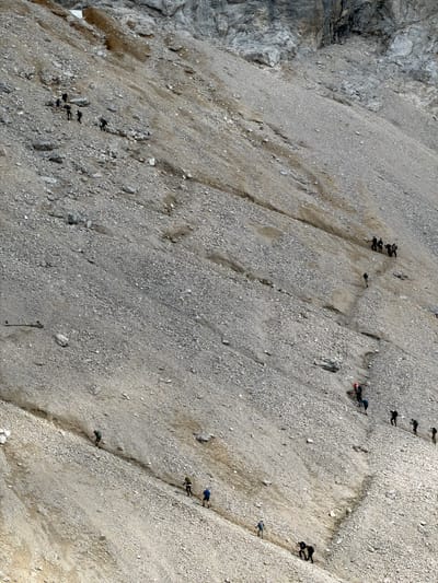 The image depicts a steep, scree-covered mountainside in Wildermieming, Austria, where a group of approximately 17 hikers are ascending a narrow, worn path. The terrain is rocky and barren, with loose stones and patches of dirt. The hikers are spread out along the trail, some walking in small groups, others individually. They are dressed in outdoor clothing, and most appear to be carrying backpacks. The path is indistinct in places, winding its way up the incline. The overall impression is one of a challenging but navigable mountain trail.
