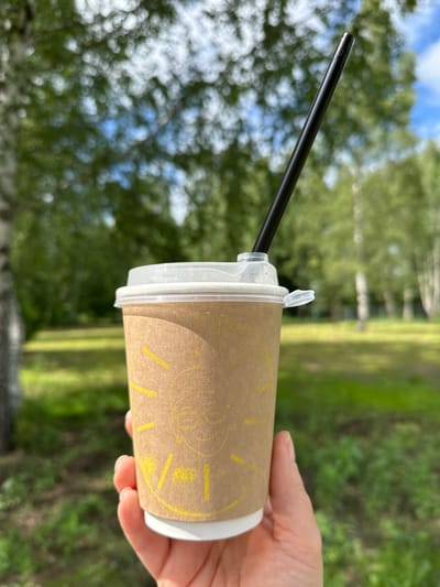 A hand holds a disposable paper cup with a lid and a black straw in an outdoor setting. The cup is brown with a yellow sun graphic featuring a face. The background is blurred and appears to be a park or wooded area with green grass and trees. The sky is visible in the upper right corner, showing some blue sky and white clouds. The location is specified as Valozhynsky Sel'savyet, Belarus.