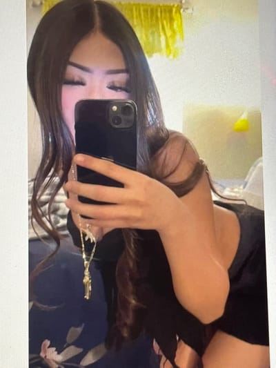 A young woman with long, dark brown hair is taking a mirror selfie. She's holding a black iPhone, partially obscuring her face. Her makeup is visible, featuring defined eyebrows and long eyelashes. She's wearing a dark top, possibly black satin shorts and a black top, and a delicate gold necklace is visible. The background shows a gold, possibly foil-wrapped item, and a neutral-colored wall. The photo is taken at an angle.