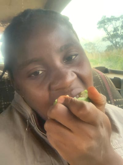 An individual with dark skin and braided hair is depicted in a close-up, angled shot, consuming a light green fruit or vegetable held in their right hand. A bite has been taken from the object. The person is wearing a light-colored zippered garment. A patterned fabric, possibly vehicle upholstery, is visible behind the individual's right shoulder. Through a window in the background, distant trees are discernible against a bright, hazy sky. The scene occurs within a vehicle in Gidan Jatau, Nigeria.