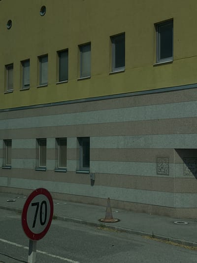 The image depicts a section of a two-story building with a yellow upper facade and a horizontally striped lower facade made of gray and beige stone. The upper facade features several rectangular windows with blinds, and two circular windows near the top. The lower facade has a row of rectangular windows. In the foreground, on a sidewalk, there is a speed limit sign indicating "70" within a red circle, and a traffic cone. The scene is set in a city environment, possibly Berg, Austria, given the building's architecture and the speed limit sign. The lighting suggests it is daytime.