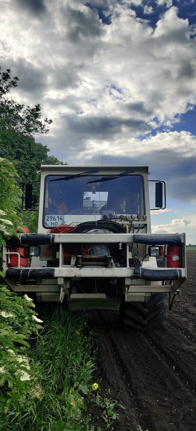 The image depicts an agricultural sprayer in a field near Kovalivs'ke, Ukraine. The machine is predominantly white, with a cabin, a set of large tires, and a visible license plate reading "29614 HO". A red fire extinguisher is mounted on the sprayer's frame. The sprayer is parked adjacent to a field with dark soil, and the foreground is filled with green vegetation and small white flowers. Above the sprayer, the sky is partly cloudy.