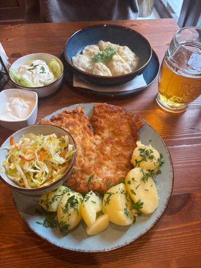 The scene depicts an indoor dining setting, likely a restaurant, within Wojnowo, Poland. A wooden table is laid with a meal for at least one person, whose upper body is partially visible in the blurred background.
The primary subject is a meal arranged on a large light blue plate. This plate holds two golden-brown, breaded cutlets, several boiled potato halves garnished with chopped parsley, and a side bowl of shredded cabbage and carrot salad.
Accompanying the main plate are additional food items and beverages:
- To the upper left, a small white bowl contains a white creamy dip.
- Adjacent to it, another small off-white bowl holds a white sauce with sliced cucumbers and a garnish of herbs.
- Centrally positioned above the main plate, a dark blue bowl contains multiple pierogi (dumplings), topped with a light sauce and fresh dill.
- To the right, a clear glass stein is filled with a light amber liquid consistent with beer, topped with a layer of foam.
The environment is characterized by the prominent wooden table surface. Natural light appears to emanate from a window in the blurred background.