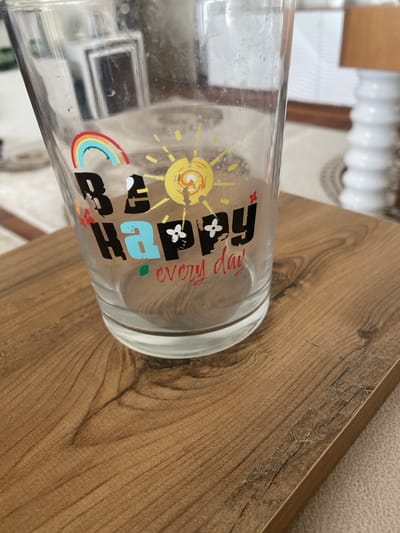 A close-up shot shows a glass with the words "BE HAPPY EVERY DAY" printed on it. The glass is sitting on a wooden surface. The background is out of focus, but appears to be an indoor setting with a white ribbed lamp and a patterned rug. The image was taken in Talas, Turkey.