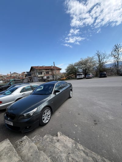 A medium-angle shot depicts a gray BMW sedan parked on an asphalt street in Sveti Konstantin, Bulgaria. The vehicle is adjacent to a concrete curb and other parked cars, including a silver sedan. Buildings with tiled roofs are visible in the background, along with trees and a blue sky with scattered clouds.