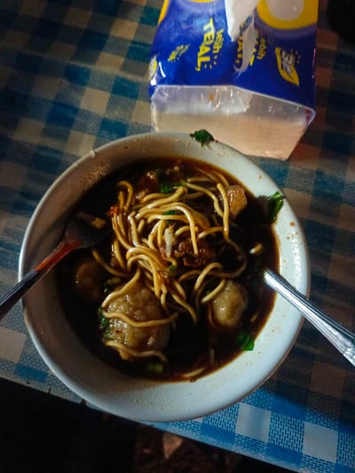 In Luwoo, Indonesia, a close-up shot captures a bowl of noodles with meatballs on a blue and white checkered tablecloth. A packet of tissues is visible in the upper right corner. Two spoons are placed in the bowl, and a fork is partially visible on the left. The food appears to be a local delicacy.