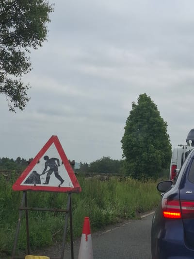 The image depicts a roadside scene on a cloudy day. In the foreground, a triangular warning sign with a red border indicates road work, showing a silhouette of a person digging. The sign is mounted on a metal stand. To the right of the sign, a traffic cone is visible. Further to the right, the rear of a dark blue car is shown, with its brake lights illuminated. Behind the car, a white van is partially visible. The background consists of a grassy verge, trees, and a stone wall. The image is taken from a low angle, suggesting the photographer is in a vehicle. There is no specific indication of the city of Barnsley, United Kingdom.