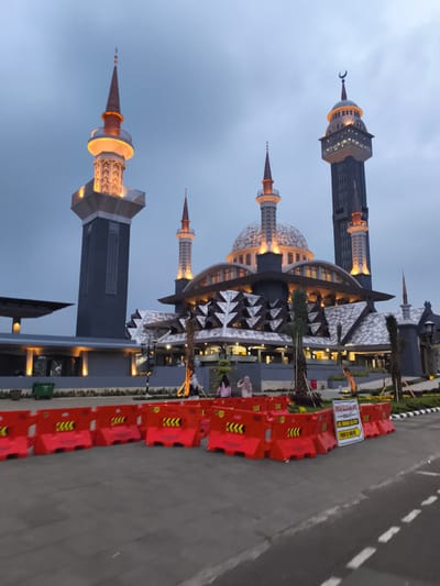 This image captures a grand, modern mosque complex in Cibinong, Indonesia, illuminated at dusk or early evening. The sky is a uniform grey-blue, suggesting an overcast day transitioning into night, with the mosque's warm artificial lights prominently on.
The main subject is the mosque itself, featuring a large central dome surrounded by several smaller domes and multiple distinct minarets. The architecture blends traditional Islamic elements with contemporary design, including intricate geometric patterns on the building's facade. Two tall minarets stand out: one on the far left with a tapering spire and an illuminated circular balcony, and another on the far right which is a very tall, dark grey structure with an elaborate, brightly lit upper section crowned with a crescent moon finial. Other smaller minarets are integrated into the main structure, also brightly lit.
In the foreground, a line of bright orange plastic road barriers, marked with yellow chevron patterns, stretches across the width of the image, separating the viewer's position from the mosque grounds. Attached to one of these barriers is a white sign with black text that reads "BISA PARKIR DI AREA INI" (Can Park In This Area) and "PARKIR DEKAT MASJID" (Parking Near Mosque), accompanied by a right-pointing arrow, indicating a nearby parking facility.
Several people are visible in the middle ground, near the mosque's entrance and on elevated walkways, appearing to be visitors or congregants. Two individuals, one in a pink shirt and another in a light-colored hijab, are clearly discernible on the left side of the main building. Green landscaping, including palm trees, surrounds the complex. A green trash bin is also visible near the base of the mosque complex on the left. The scene evokes a sense of grandeur and peaceful evening activity at a significant religious landmark.