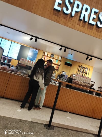A couple stands in front of a coffee shop counter. The woman is wearing a light-colored sweater and dark pants. The man is wearing a dark jacket and light-colored jeans. A long counter with a wood-paneled front runs along the space with coffee machines and supplies behind it. Above the counter, a menu board is visible, and the word "ESPRESSO" is spelled out in large white letters. A black metal pole stands in the foreground. The floor is light gray tile.