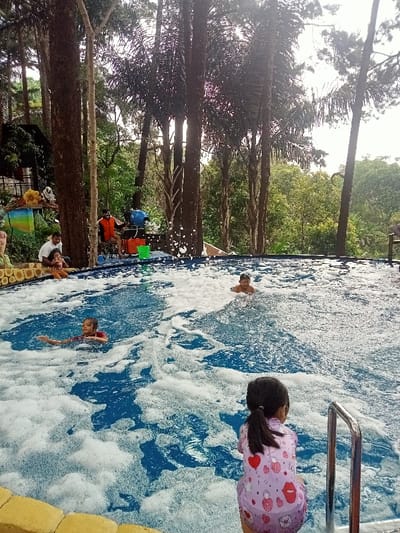 An outdoor scene in Kuwukan, Indonesia, depicts a swimming pool filled with white foam, surrounded by dense forest. In the foreground, a young girl wearing a light purple swimsuit with heart and lock patterns sits at the pool's edge with her back to the camera, near a stainless steel handrail. Two young boys are partially submerged in the foamy blue water; one in a red shirt is on the left, and another is further right, visible from the chest up. On the left side of the pool, an adult male with a beard sits on the edge, holding a small child. In the background, two adult males are positioned near a blue foam-generating machine and two buckets (one red, one green). The entire area is heavily treed with tall, slender trees, creating a shaded environment under an overcast sky.