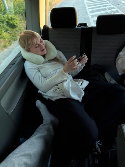 A sideways, overhead view shows a woman sleeping on a bus or train. She has blonde hair and wears a travel neck pillow. She is positioned near a window where green trees and foliage are visible. The woman wears a white long-sleeve shirt or sweater, and a light-colored sweatshirt or jacket is draped over her. She holds a pair of glasses in one hand. A person wearing dark pants and shoes is seated nearby. Another person wears a gray sweat suit and socks, also possibly sleeping. The seat, and what appears to be an arm rest, are visible. The specific location of Loznica, Serbia, cannot be confirmed with the given information.
