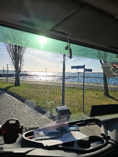 The image depicts an outdoor scene viewed from inside a vehicle, through its front windshield. The setting is the intersection of Berliner Straße and Hafenstraße in Bitterfeld-Wolfen, Germany, with the Goitzschesee visible in the background.
The scene is illuminated by bright sunlight, creating significant glare on the upper, green-tinted section of the windshield and reflecting intensely off the expansive, slightly rippled surface of the Goitzschesee. The sky above the lake is clear and blue.
In the midground, a paved road or path runs parallel to the lake. Between the road and the water lies a grassy strip dotted with numerous scattered dry, brown leaves, suggesting an autumn or early winter season. Several bare-branched deciduous trees stand along this strip, accompanied by multiple dark, vertical poles, which appear to be streetlights or structural supports. A dark-colored barrier or railing is visible along the water's edge. A street sign at the intersection clearly displays "Berliner Straße" pointing left and "Hafen Str." pointing right.
The foreground details the interior of the vehicle, specifically the dark-colored dashboard. On the left side of the dashboard, a dark-colored phone holder or mount is present. Centrally, a stack of papers and documents, some with blue or metallic strips, rests on the dashboard surface. To their right, a dark, circular dashboard organizer with apparent cup holders is visible. Further right, a small, light-colored rectangular device is affixed to the dashboard. A grey sun visor extends across the top of the frame. A small, dark, circular object hangs from the sun visor, suspended by a string and secured to the windshield with a suction cup. No individuals are visible in the scene. The vehicle appears stationary.