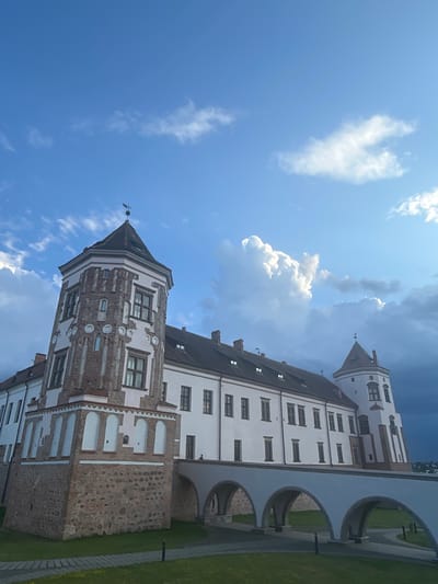 The image shows the Mir Castle Complex, a UNESCO World Heritage site located in the village of Mir, Karelichy District, Grodno Region, Belarus. The castle, viewed from a low angle, features a prominent gothic tower on the left, constructed with red brick and white plaster accents, and a longer, white-plastered building extending to the right, with multiple windows and a tiled roof. A white arched bridge connects the castle to what appears to be a fortified wall or another part of the complex. The sky above is a vibrant blue with scattered white cumulus clouds, some of which are illuminated by sunlight, while darker, stormier clouds are visible in the far right background. The foreground consists of a well-maintained green lawn with paved walkways. There are no people visible in the image. The overall scene depicts a historic architectural landmark under a dynamic sky.