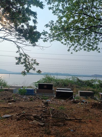 The image shows a view overlooking a body of water in Kothacheruvu, India. In the foreground, there is a section of disturbed earth covered with small stones, branches, and litter. A series of concrete and stone burial markers or mausoleums are situated on a slightly raised platform, interspersed with scrub vegetation. The middle ground displays the water body with distant hills faintly visible on the horizon. Overhead power lines run across the scene. Trees are present at the top of the frame, partially obscuring the sky. The scene appears to be a cemetery or burial ground situated near a lake or river.