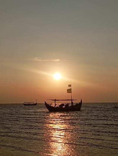 The image displays a maritime scene at either sunrise or sunset over the waters of Tritunggal, Indonesia. The sky exhibits a gradient from a pale grey-blue at the top to a bright orange-yellow at the horizon, with the sun appearing as a brilliant orb in the upper-middle right quadrant. The water's surface is rippled, showing a strong, shimmering reflection of the sun's orange-yellow light extending towards the viewer.
A large, dark-silhouetted traditional wooden fishing boat occupies the mid-ground, positioned slightly right of the frame's center. This vessel features distinctively high, curved bow and stern sections. A central structure, possibly a canopy or cabin, is visible, and a mast rises from it, flying two rectangular flags: one light-colored (beige/yellow) and one darker-colored (brown/dark yellow). Indistinct forms consistent with occupants or gear are discernible within the boat's dark silhouette. In the distant left background, a second, smaller, similarly silhouetted traditional fishing boat is visible on the water. No specific human actions or interactions are clearly identifiable due to the silhouettes. The environment is calm.
