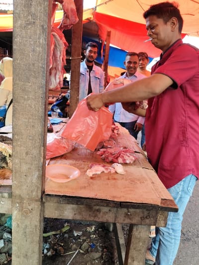 The image depicts an outdoor market stall in Tuha Gp Gajah, Indonesia. In the immediate foreground to the right, a man in a red collared shirt and blue jeans is actively handling cuts of raw meat on a weathered wooden table. A lit cigarette is positioned in his mouth. His left hand holds a red plastic bag open, while his right hand holds a piece of light-colored meat, appearing to place it into the bag or onto the table. The wooden table surface is covered with a light-colored, stained material and displays various pieces of red and lighter-colored raw meat. A small, light orange plastic bowl containing liquid is situated on the table to the left.
Behind the man handling the meat, three other men are visible. From left to right: a man with a beard wearing a light blue denim jacket, a smiling man in a light blue button-up shirt, and a partially visible man in an orange t-shirt. Further in the background, hanging pieces of raw meat are suspended from wooden beams. The stall structure consists of visible wooden pillars and beams, partially covered by red and blue tarpaulin canopies. To the far left, a person wearing a light-colored head covering and yellow garment is partially visible. The ground beneath the wooden table is unpaved, showing dirt and scattered debris including plastic fragments.