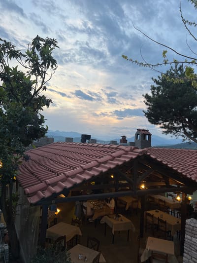 The image captures an outdoor dining area in Kastraki, Greece, at twilight. The focus is on a restaurant's red-tiled roof and wooden structure, under which tables are set for dining. Light fixtures illuminate the tables. Beyond the restaurant, the landscape features a medieval stone tower and rocky cliffs against a backdrop of a cloudy sky. Vegetation, including trees and branches, frames the scene in the foreground and top left corner.